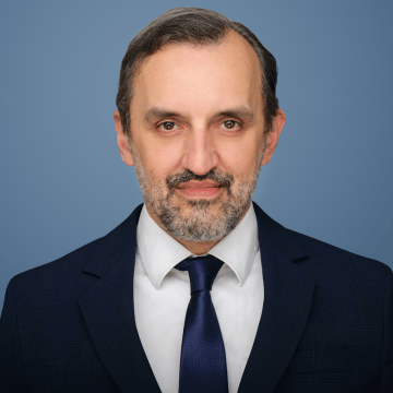 DR. NENAD ANTIC is posing for a professional headshot in a suit and tie against a blue background